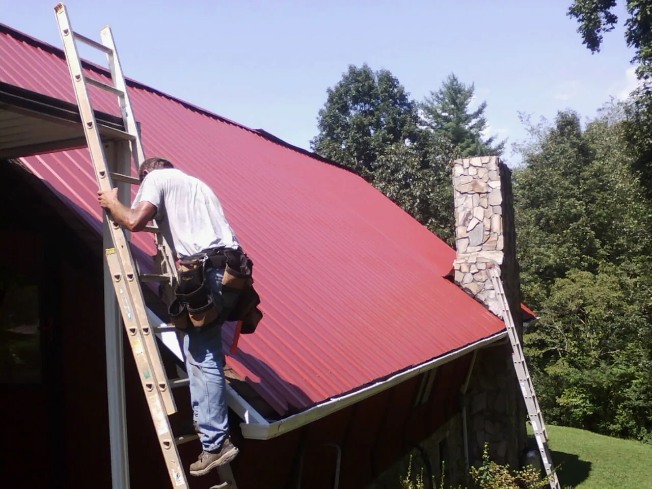 Roofer on a ladder beside a red metal roof and stone chimney — Soffit & Fascia Repair by Roofing Brothers in Sparrows Point